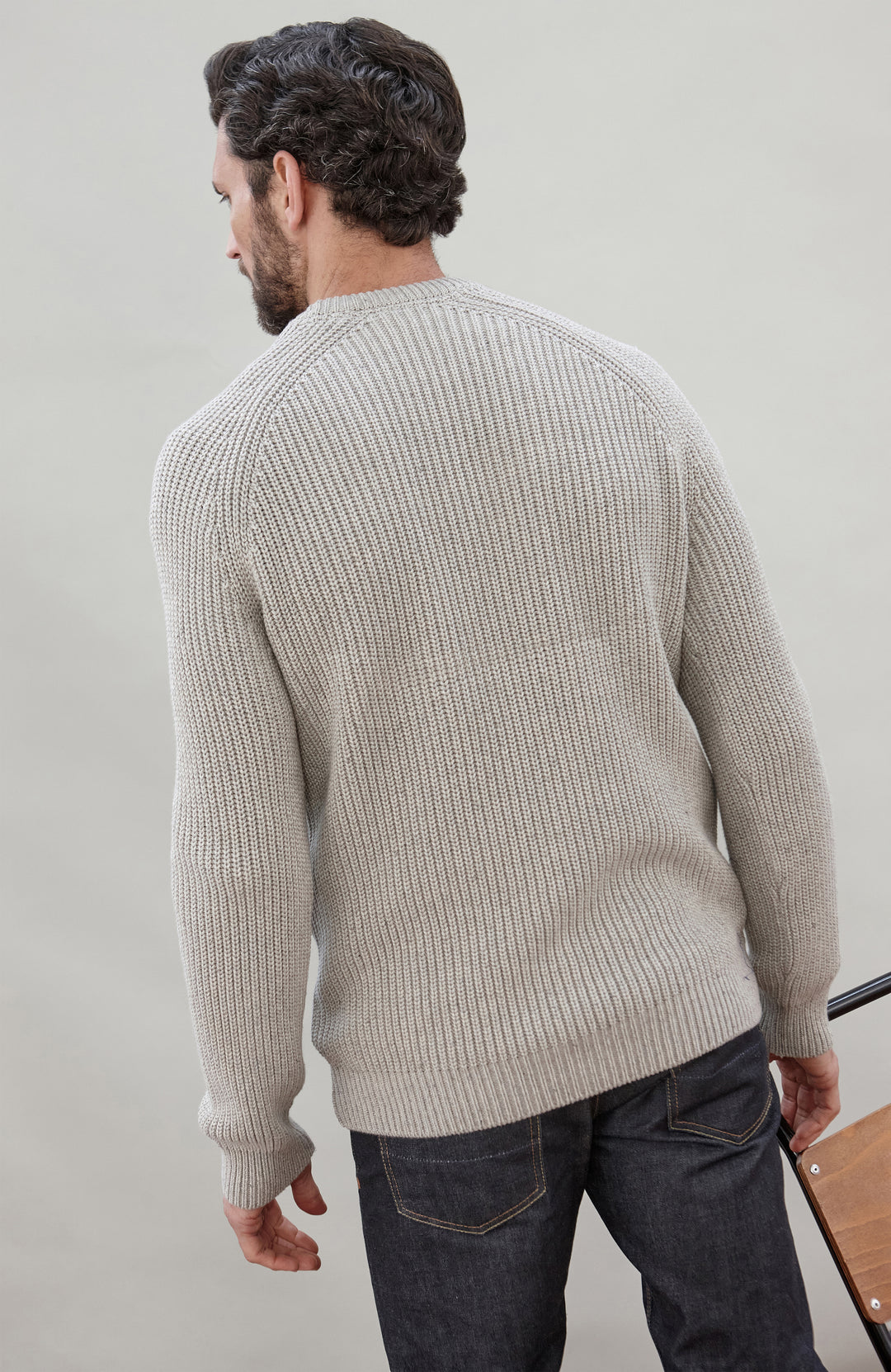 Man sitting on a chair wearing a light gray cotton fisherman sweater by Paul James Knitwear and dark jeans against a plain background
