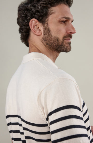 A man wearing a short-sleeve breton stripe knit polo shirt by paul james knitwear with horizontal black stripes on an off-white base, paired with dark trousers, against a solid light gray background.