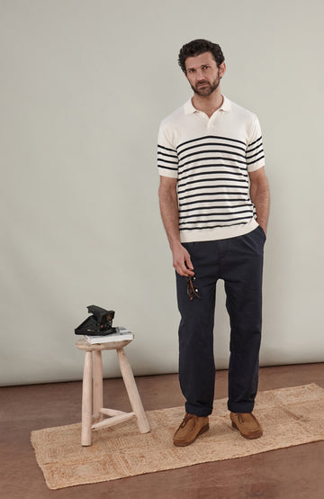 A man wearing a short-sleeve breton stripe knit polo shirt by paul james knitwear with horizontal black stripes on an off-white base, paired with dark trousers, against a solid light gray background.