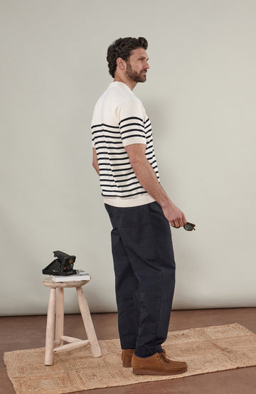 A man wearing a short-sleeve breton stripe knit polo shirt by paul james knitwear with horizontal black stripes on an off-white base, paired with dark trousers, against a solid light gray background.