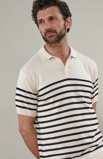 A man wearing a short-sleeve breton stripe knit polo shirt by paul james knitwear with horizontal black stripes on an off-white base, paired with dark trousers, against a solid light gray background.
