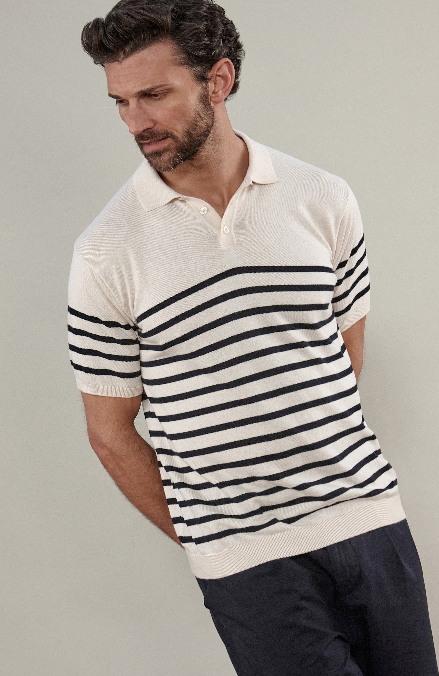 A man wearing a short-sleeve breton stripe knit polo shirt by paul james knitwear with horizontal black stripes on an off-white base, paired with dark trousers, against a solid light gray background.