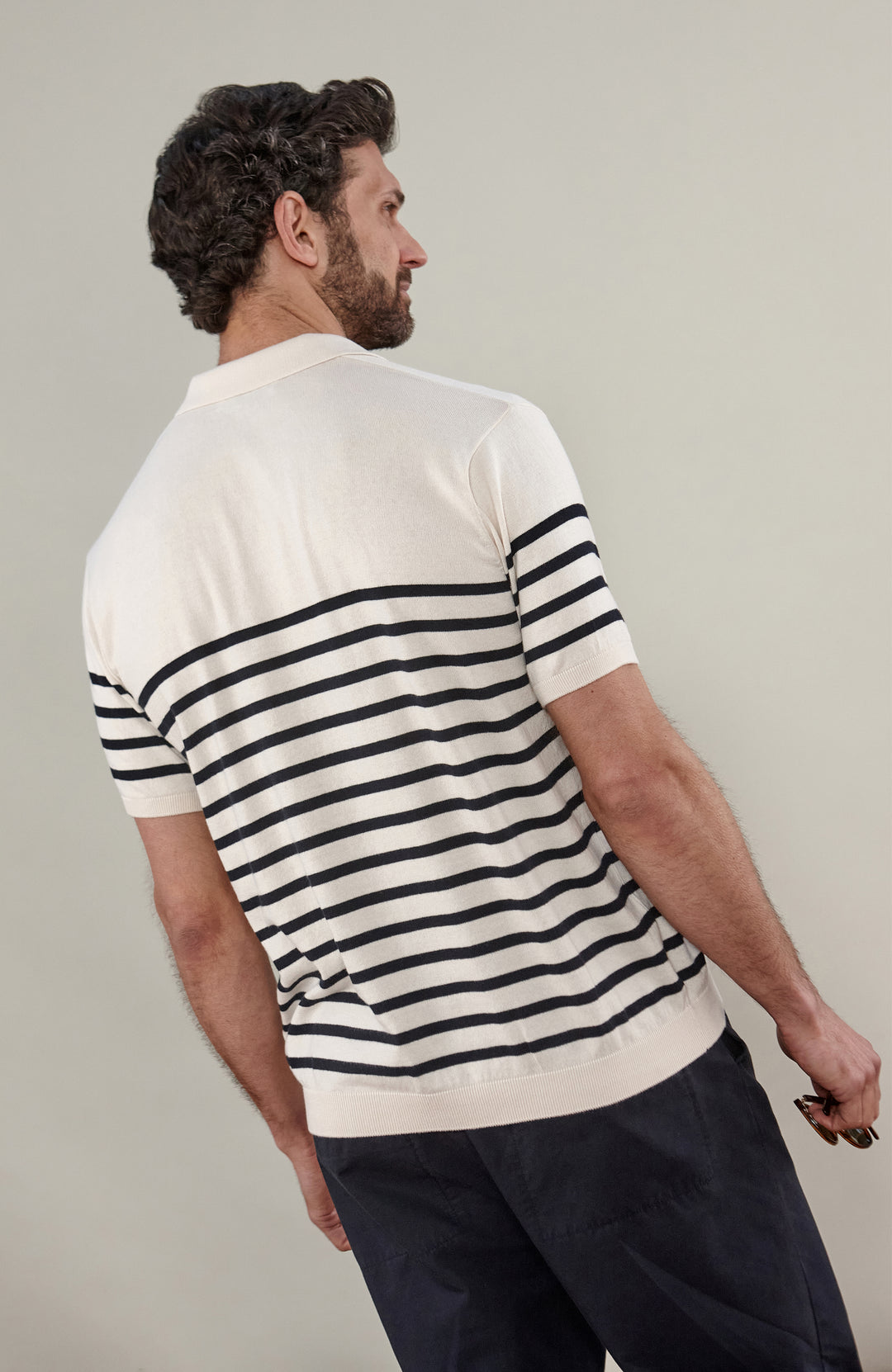 A man wearing a short-sleeve breton stripe knit polo shirt by paul james knitwear with horizontal black stripes on an off-white base, paired with dark trousers, against a solid light gray background.