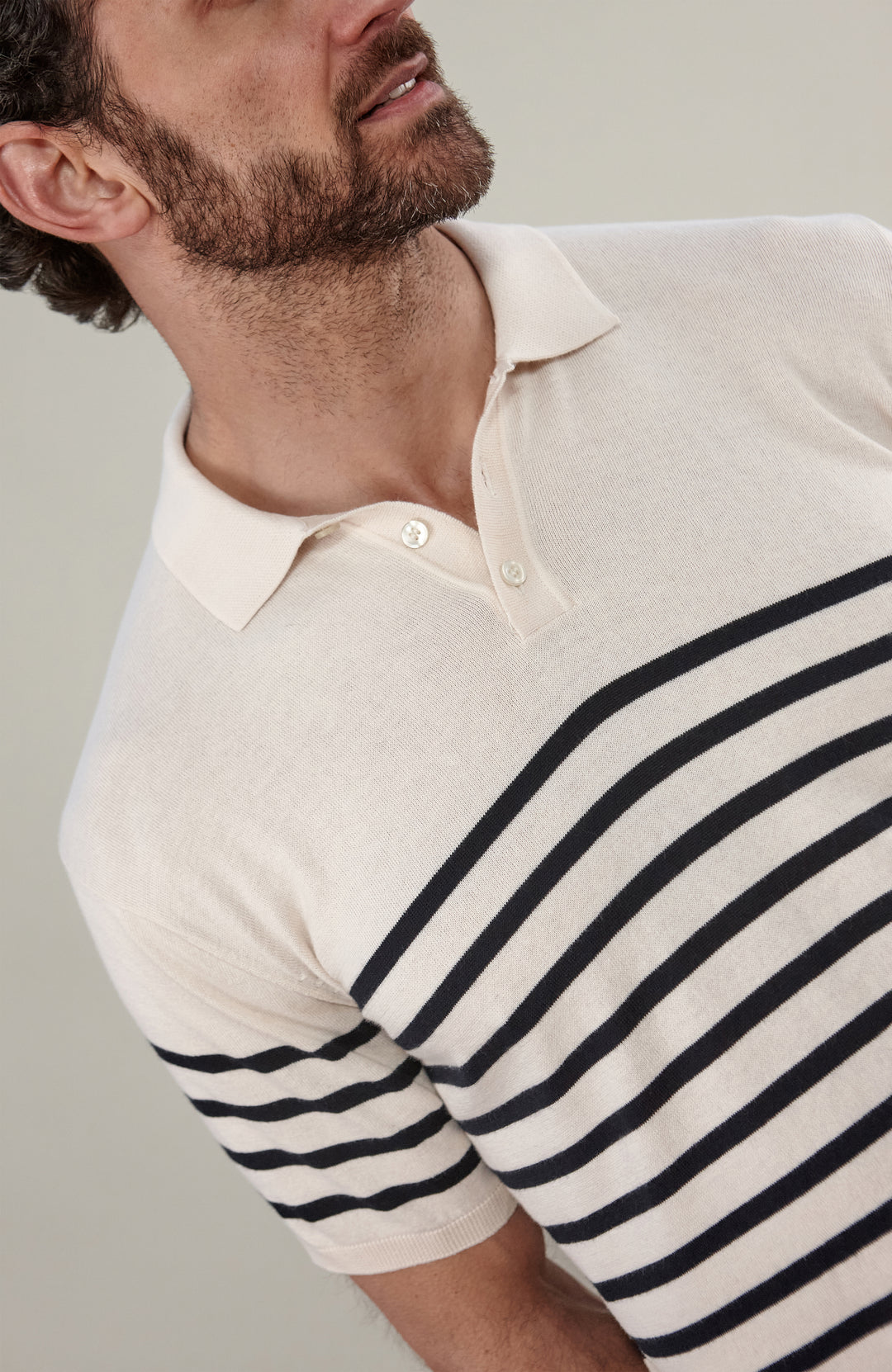 A man wearing a short-sleeve breton stripe knit polo shirt by paul james knitwear with horizontal black stripes on an off-white base, paired with dark trousers, against a solid light gray background.