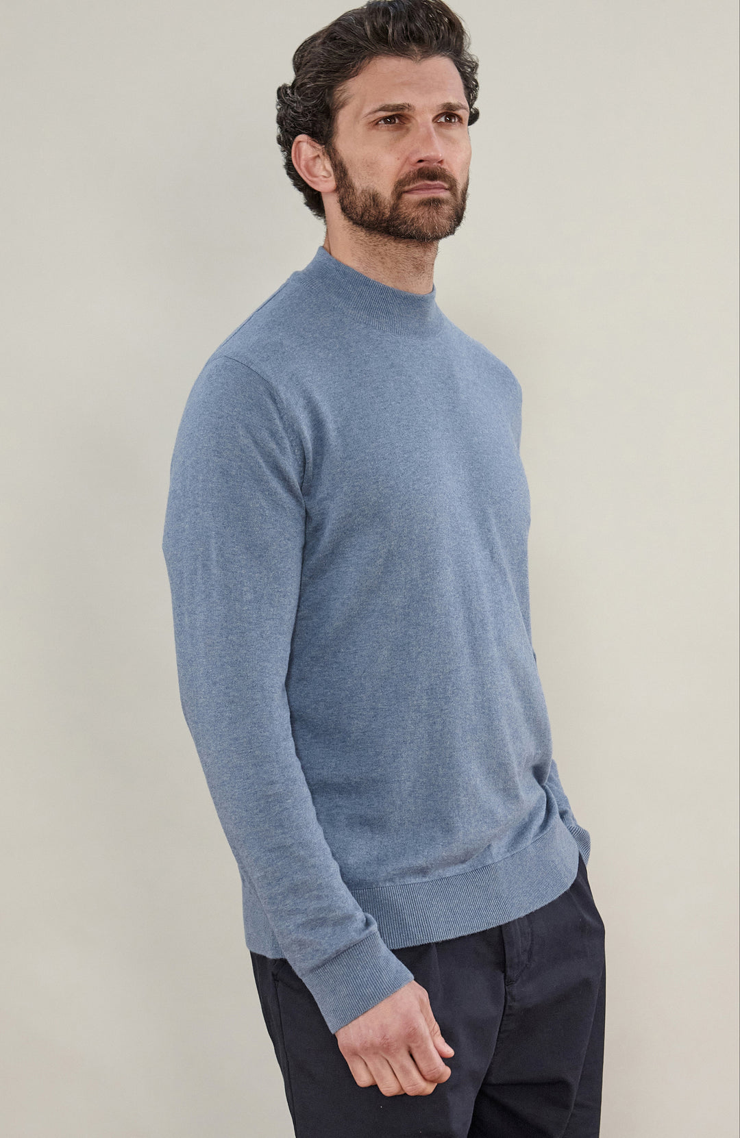 A man wearing a light blue knit mock neck sweater by Paul James Knitwear and dark trousers, standing against a plain light beige studio background.