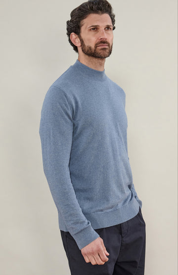 A man wearing a light blue knit mock neck sweater by Paul James Knitwear and dark trousers, standing against a plain light beige studio background.