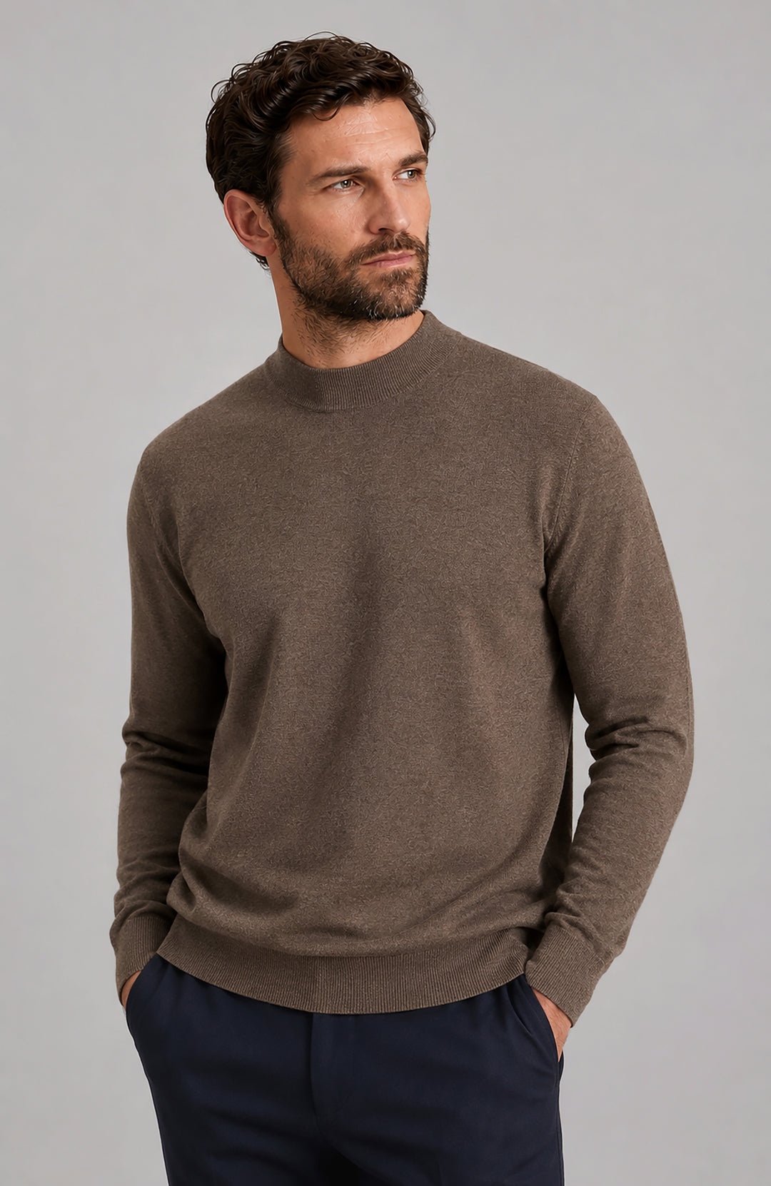 A man wearing a brown long-sleeve knit mock neck sweater by Paul James Knitwear and navy trousers, standing against a solid gray studio background.