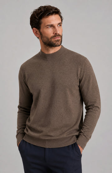 A man wearing a brown long-sleeve knit mock neck sweater by Paul James Knitwear and navy trousers, standing against a solid gray studio background.