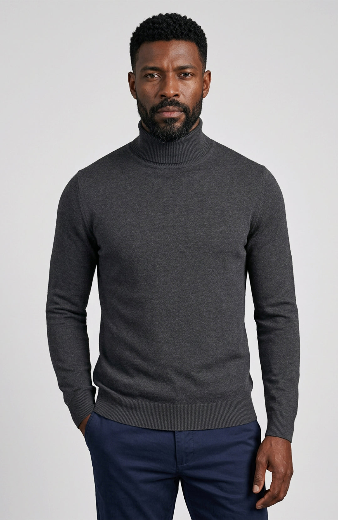 A man wearing a dark gray heathered turtleneck sweater Paul James Knitwear and navy blue trousers, standing against a light gray studio background.
