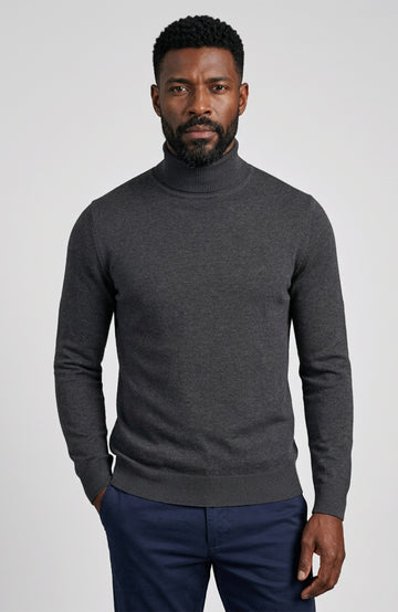 A man wearing a dark gray heathered turtleneck sweater Paul James Knitwear and navy blue trousers, standing against a light gray studio background.