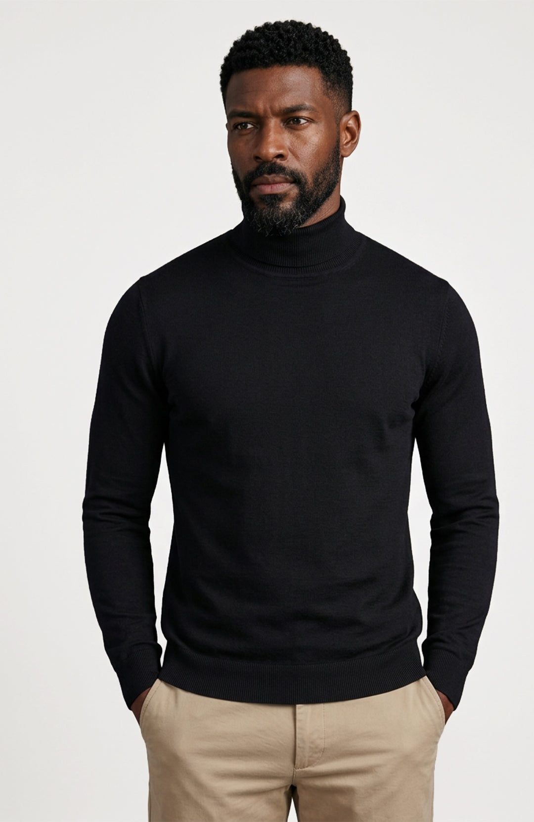 A man wearing a black long-sleeve cotton turtleneck sweater by Paul James Knitwear and beige trousers, standing against a light gray studio background.