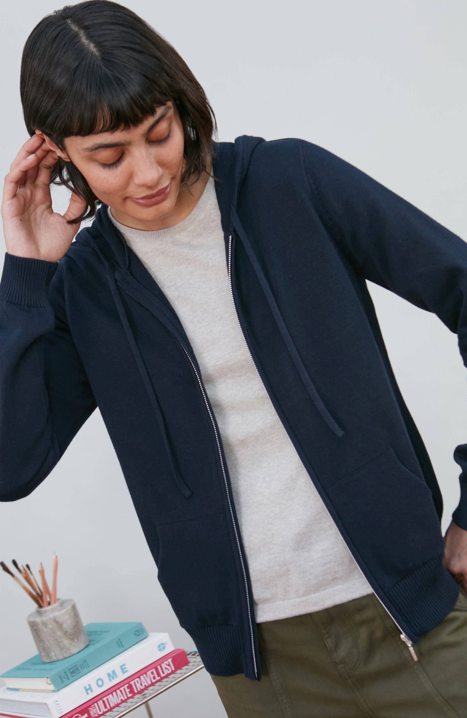 A woman wearing a navy blue zip-up hoodie by Paul James Knitwear over a light beige crewneck sweater and olive green pants, standing next to a small stack of books on a light grey background.
