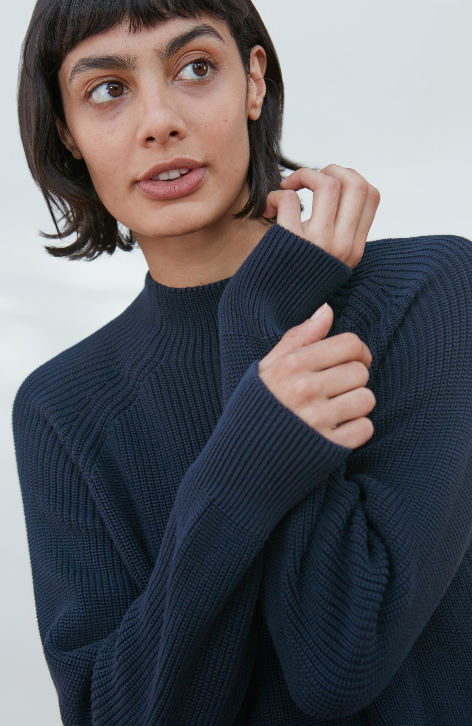 A close-up of a woman wearing a dark navy ribbed knit mock-neck sweater by Paul James Knitwear, looking off-camera with her hand near her collarbone.
