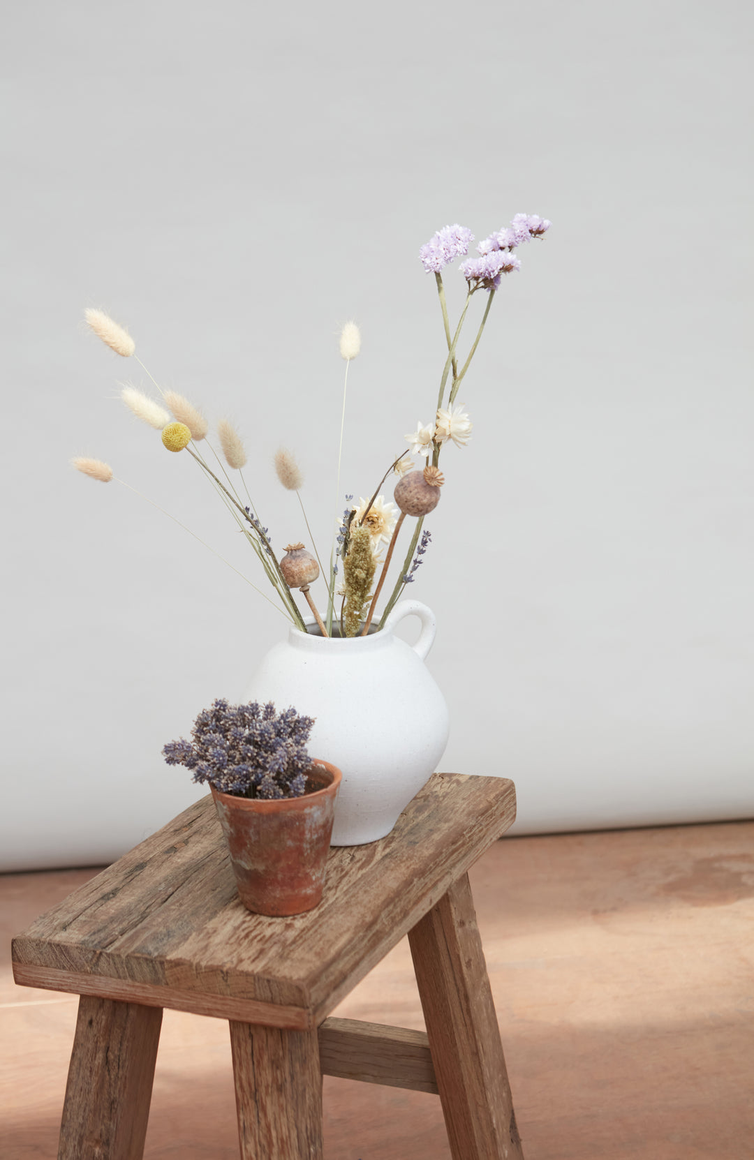White vase with dried flowers on a wooden stool against a plain background by Paul James Knitwear