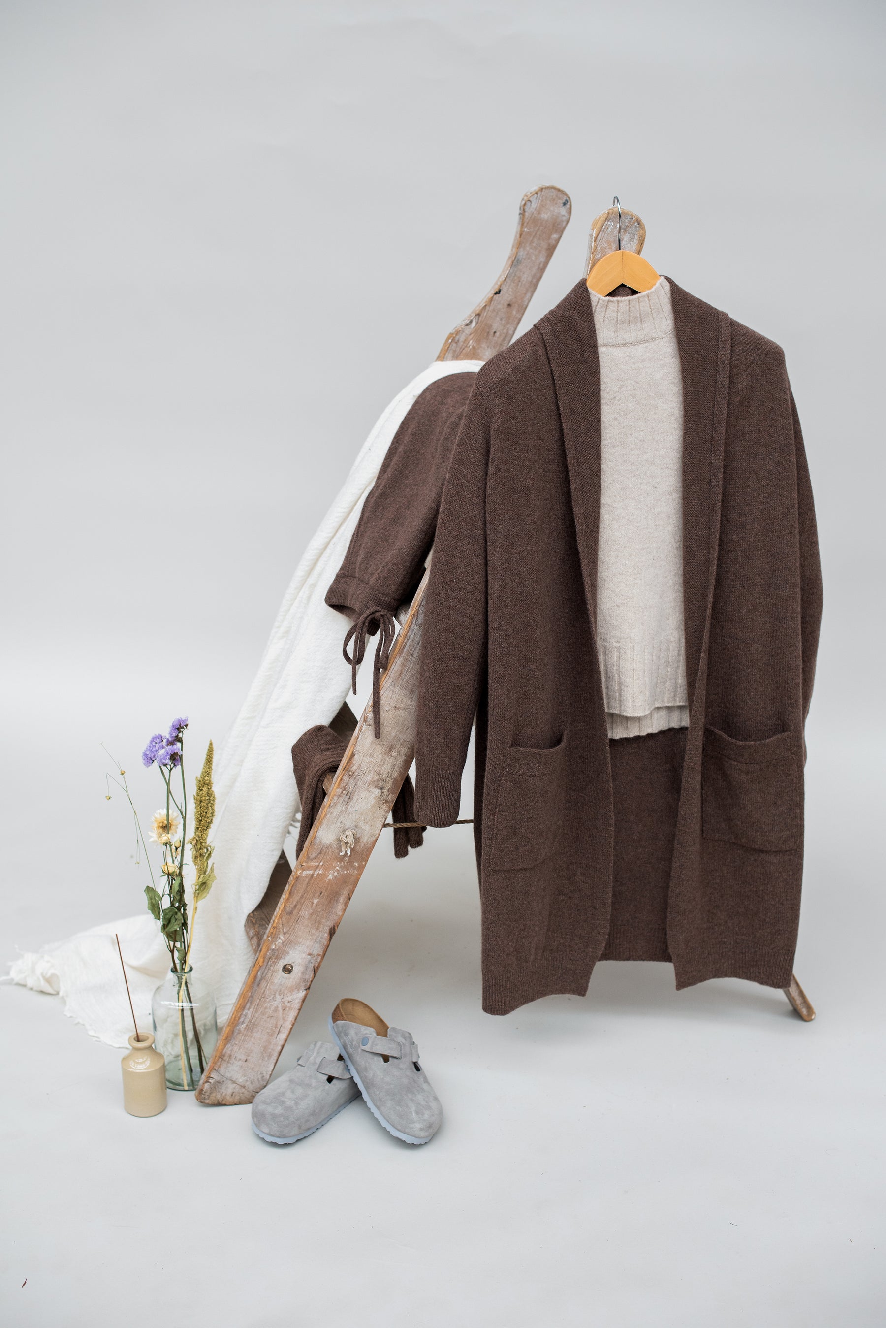 Brown wool cardigan, white shirt, gray slippers, and wooden shoes on a white background