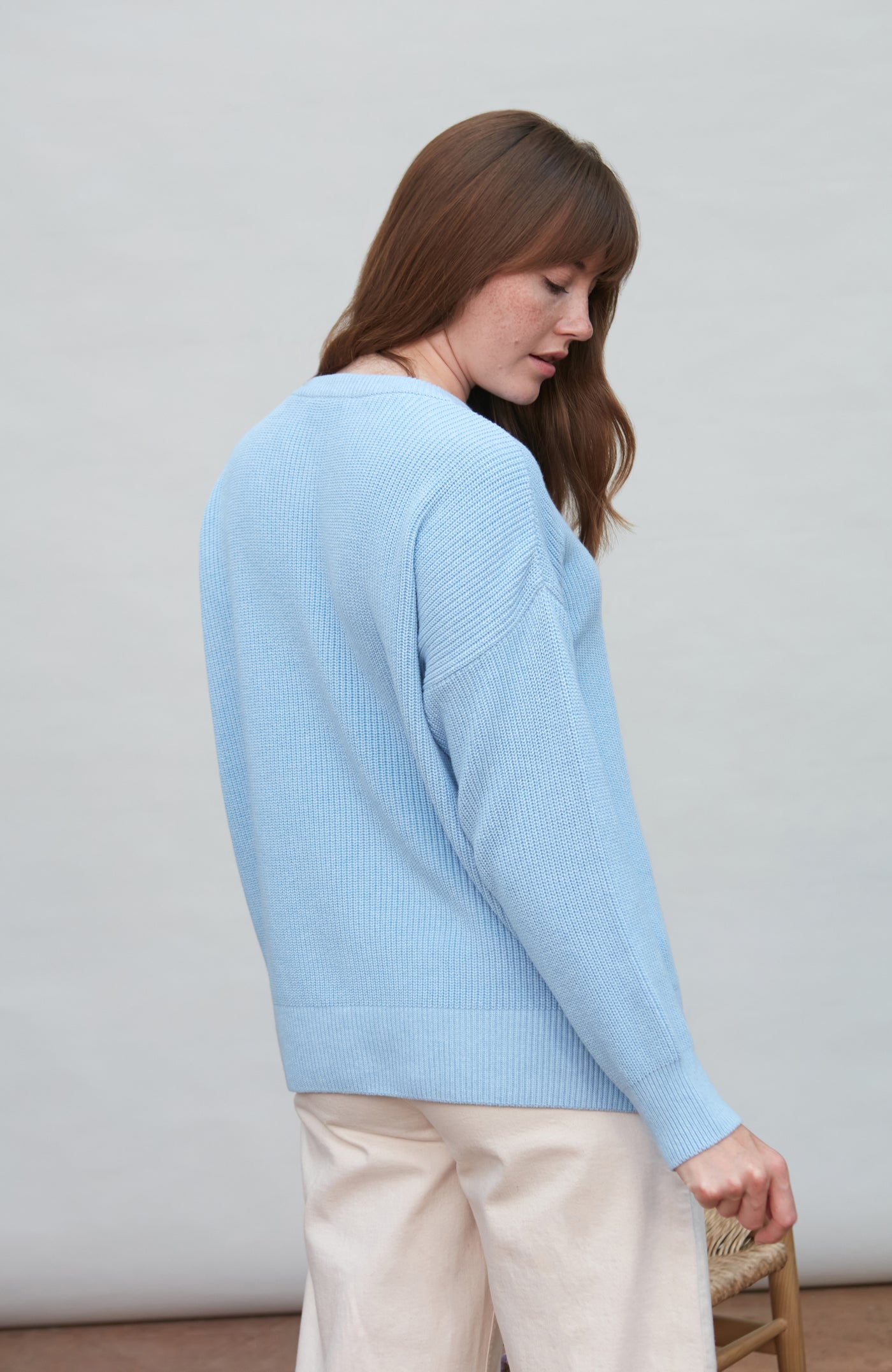 Woman wearing a light blue cotton fisherman rib sweater and beige pants against a plain background