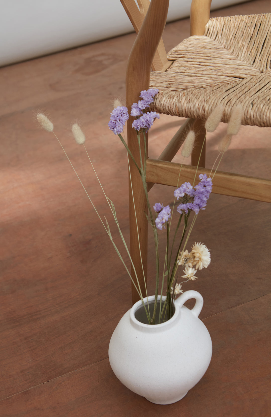 White vase with dried flowers on a wooden floor next to a wicker chair