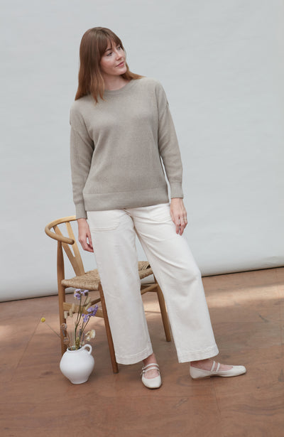 Woman wearing a beige cotton fisherman rib sweater and white pants standing in a minimalistic room.