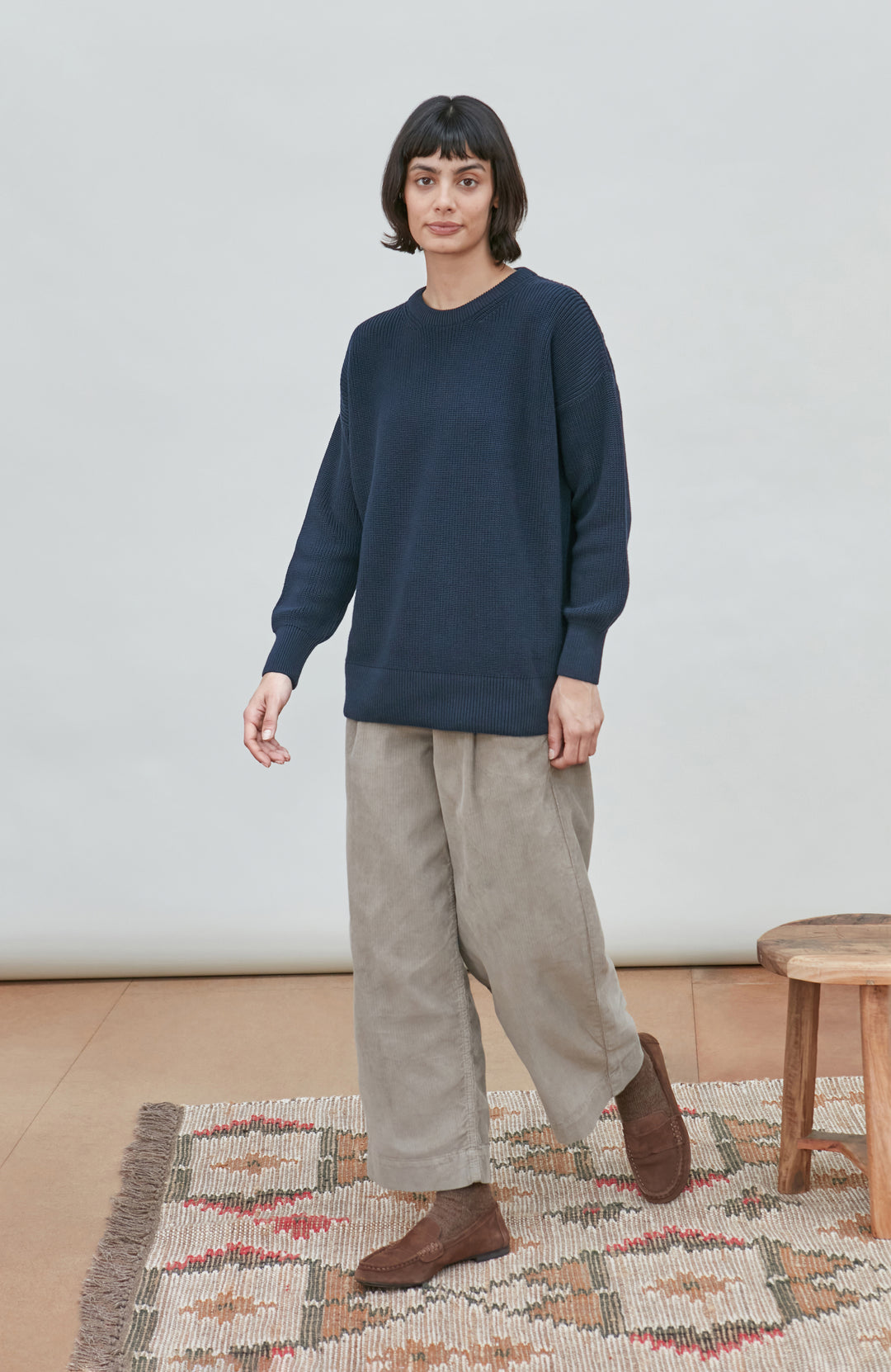 A woman stands in a minimalist room wearing a dark navy crewneck fisherman sweater by Paul James Knitwear, loose light beige trousers, and brown boots, with a patterned rug and a small wooden stool visible.