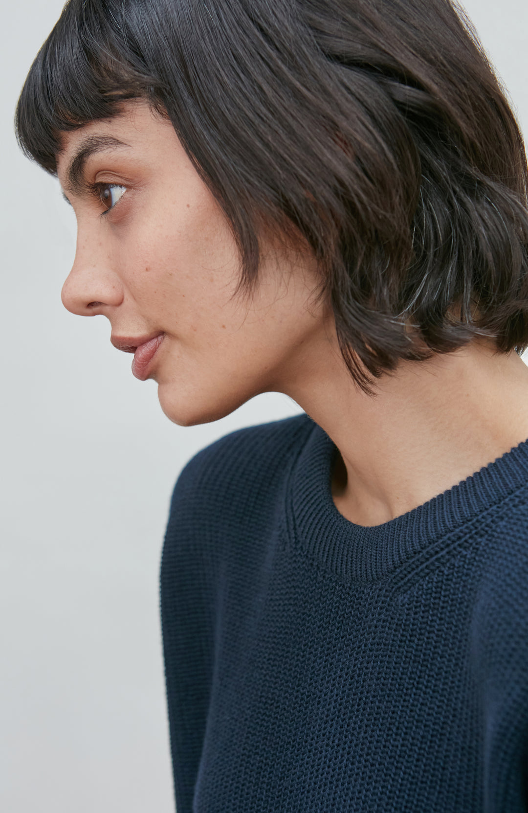 A woman wearing a dark navy blue knit crewneck fisherman sweater by paul james knitwear and beige trousers, standing against a light gray studio background.