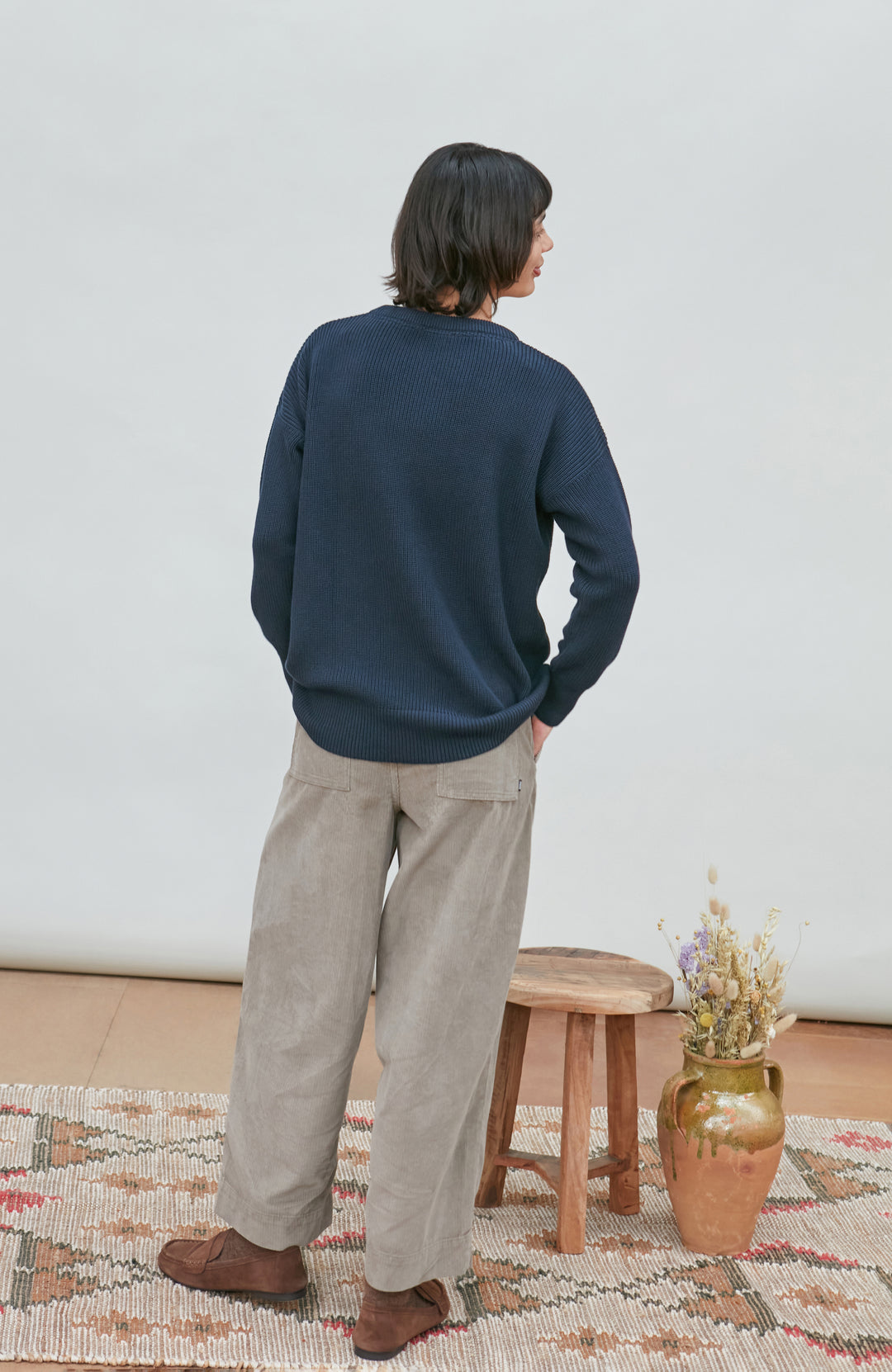 A woman wearing a dark navy blue knit crewneck fisherman sweater by paul james knitwear and beige trousers, standing against a light gray studio background.