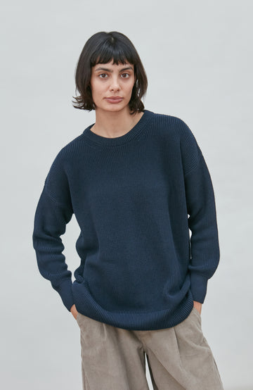 A woman wearing a dark navy blue knit crewneck fisherman sweater by paul james knitwear and beige trousers, standing against a light gray studio background.