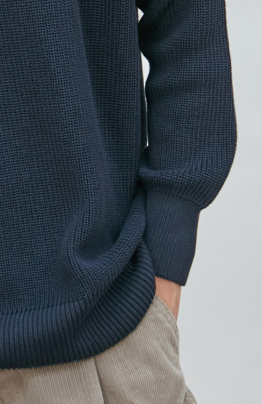 A woman wearing a dark navy blue knit crewneck fisherman sweater by paul james knitwear and beige trousers, standing against a light gray studio background.