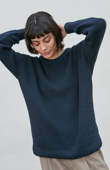 A woman wearing a dark navy blue ribbed cotton fisherman knit sweater by Paul James Knitwear, posing with her hands behind her head against a plain light gray background.