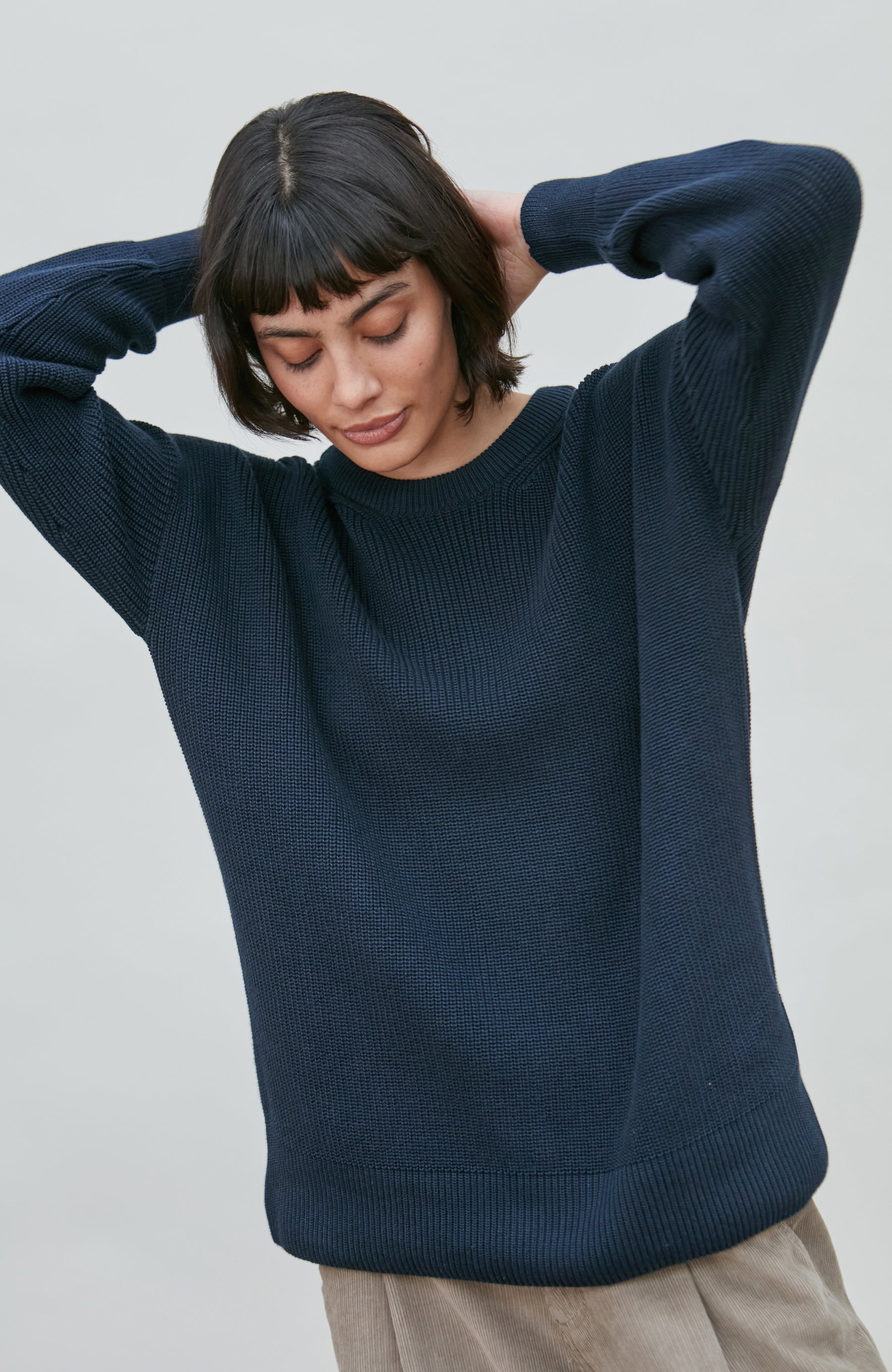 A woman wearing a dark navy blue ribbed cotton fisherman knit sweater by Paul James Knitwear, posing with her hands behind her head against a plain light gray background.
