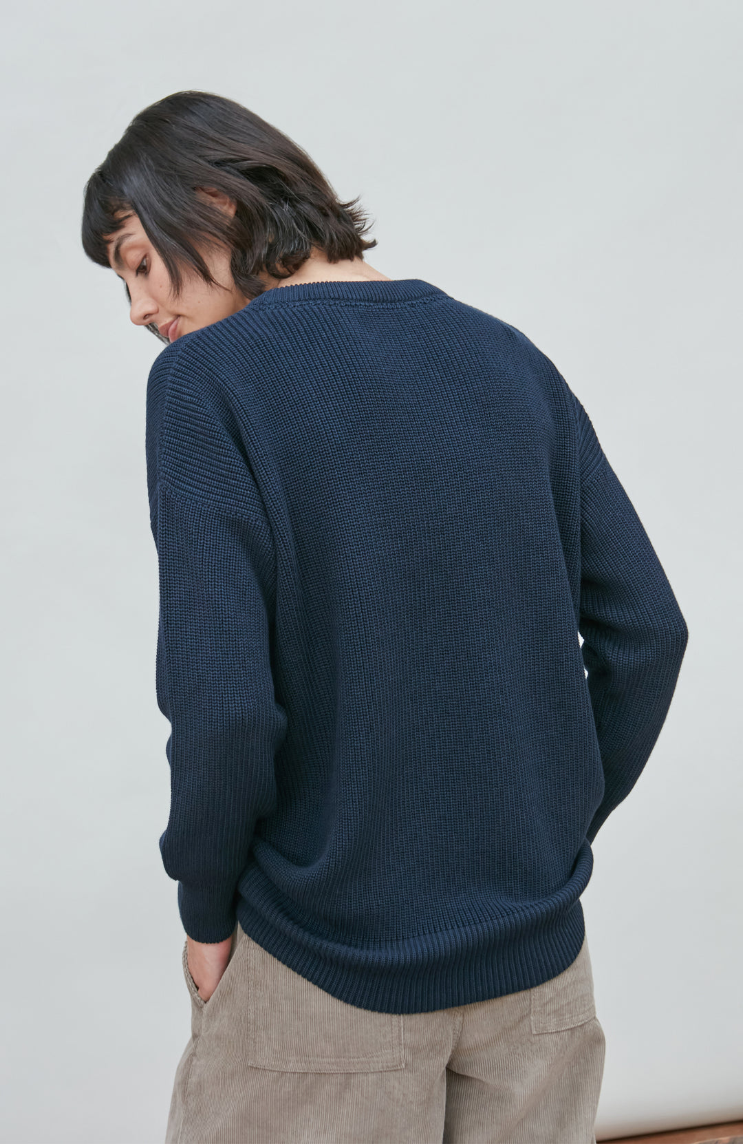 A woman wearing a dark navy blue knit crewneck fisherman sweater by paul james knitwear and beige trousers, standing against a light gray studio background.