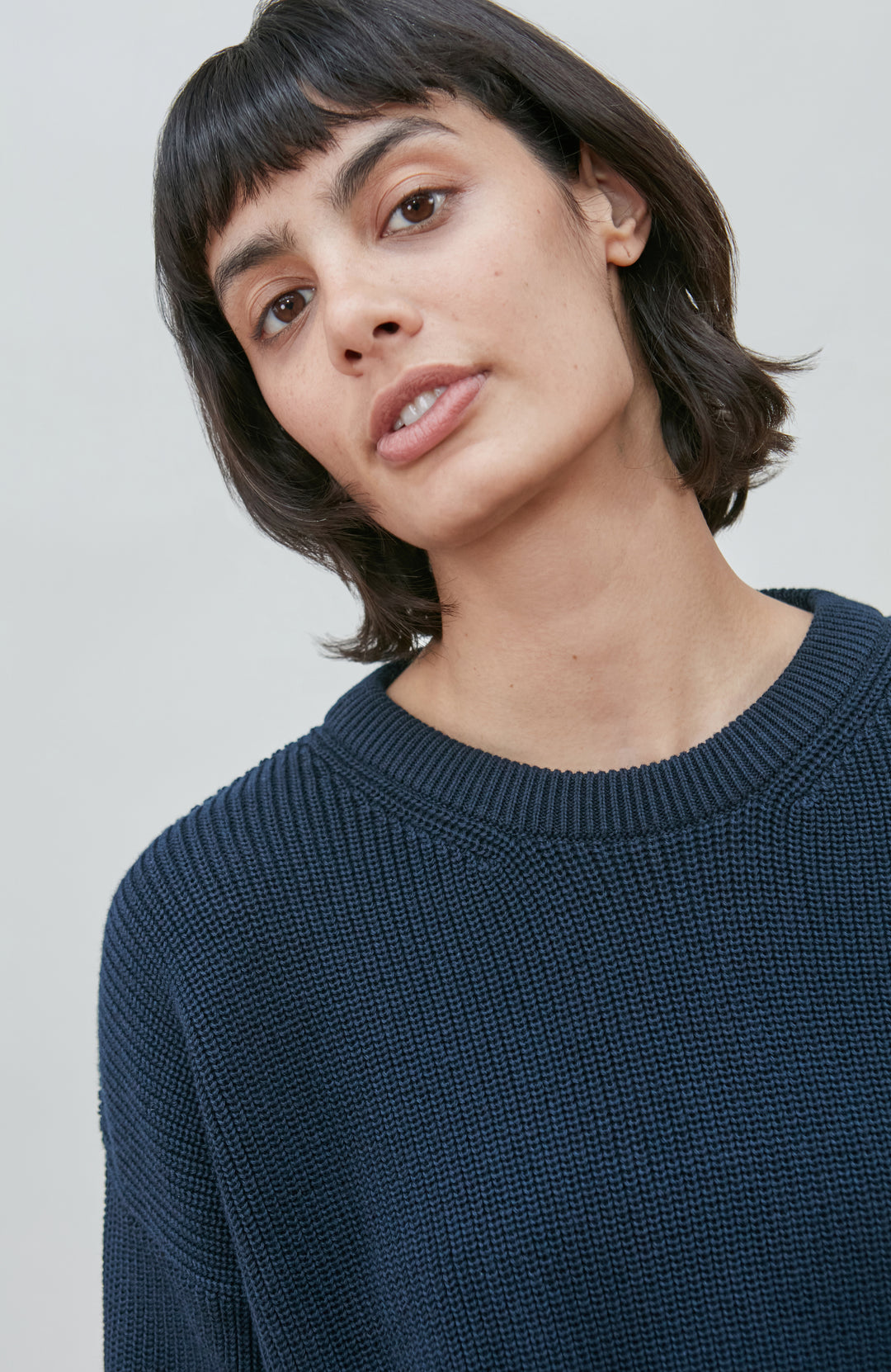 A woman wearing a dark navy blue knit crewneck fisherman sweater by paul james knitwear and beige trousers, standing against a light gray studio background.