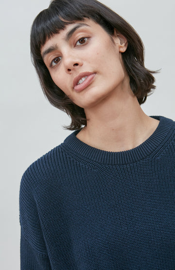 A woman wearing a dark navy blue knit crewneck fisherman sweater by paul james knitwear and beige trousers, standing against a light gray studio background.