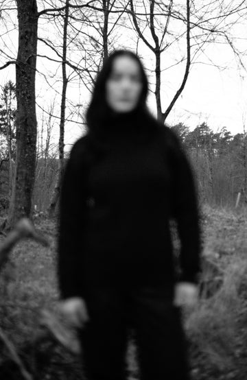 A woman stands on a fallen tree log in a wooded outdoor setting, wearing a dark navy turtleneck sweater by Paul James Knitwear, dark navy pants, and dark boots.