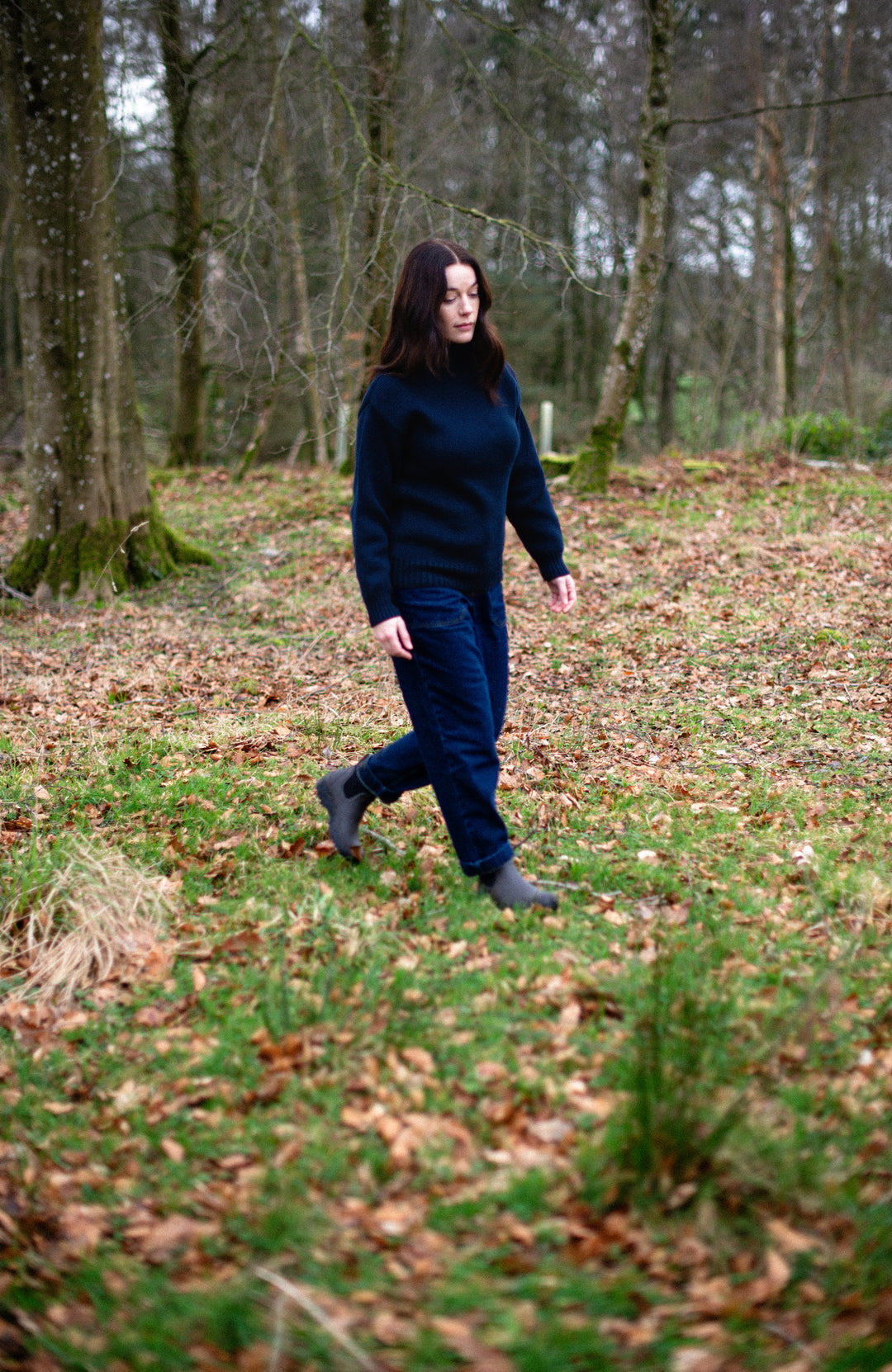 Woman in a dark blue cotton submariner roll neck jumper by Paul James Knitwear walking through a forest with bare trees and fallen leaves on the ground.