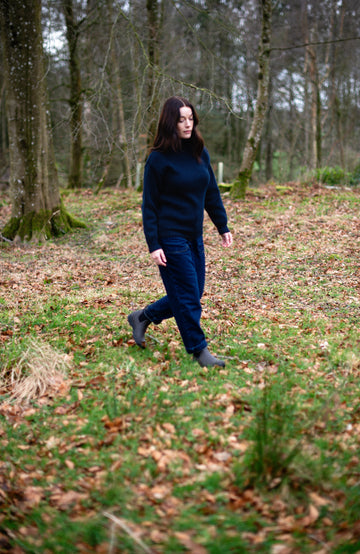 Woman in a dark blue cotton submariner roll neck jumper by Paul James Knitwear walking through a forest with bare trees and fallen leaves on the ground.