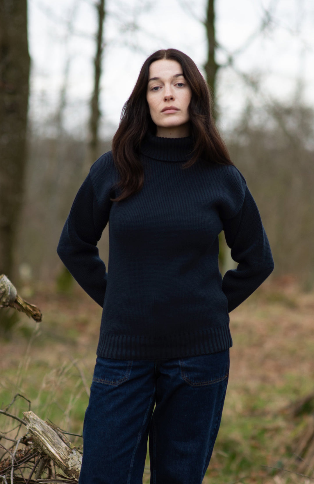 A woman stands on a fallen tree log in a wooded outdoor setting, wearing a dark navy turtleneck sweater by Paul James Knitwear, dark navy pants, and dark boots.