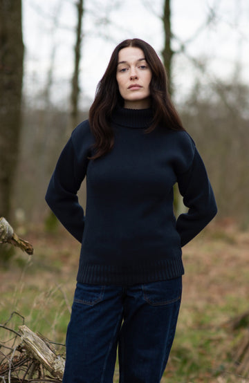 A woman stands on a fallen tree log in a wooded outdoor setting, wearing a dark navy turtleneck sweater by Paul James Knitwear, dark navy pants, and dark boots.