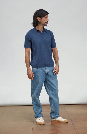 A man wearing a navy blue cotton short-sleeve polo shirt, light blue jeans, and off-white sneakers, standing against a plain light gray studio background. Made by Paul James Knitwear.