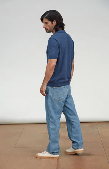 A man wearing a navy blue short-sleeve polo shirt, light blue jeans, and white sneakers, shown from a side-back angle against a plain studio background. Made by Paul James Knitwear.