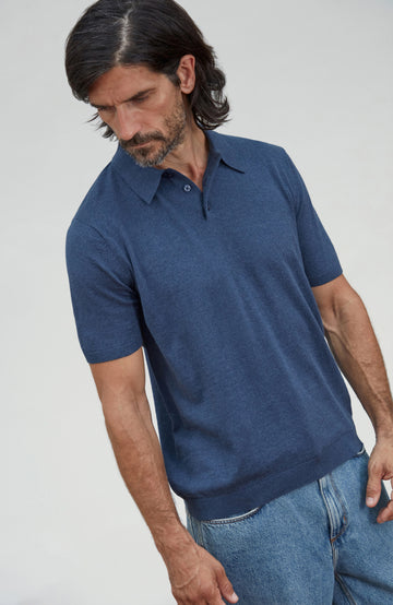 A man wearing a short-sleeve knit polo shirt in a heathered navy blue color, paired with blue jeans, photographed against a light gray studio background. Made by Paul James Knitwear.