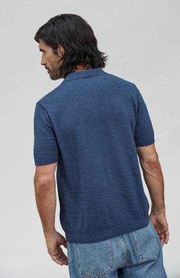 A man with long dark hair is shown from the back wearing a short-sleeve knit polo shirt in heathered navy blue, paired with light blue jeans, against a plain light gray studio background. Made by Paul James Knitwear.