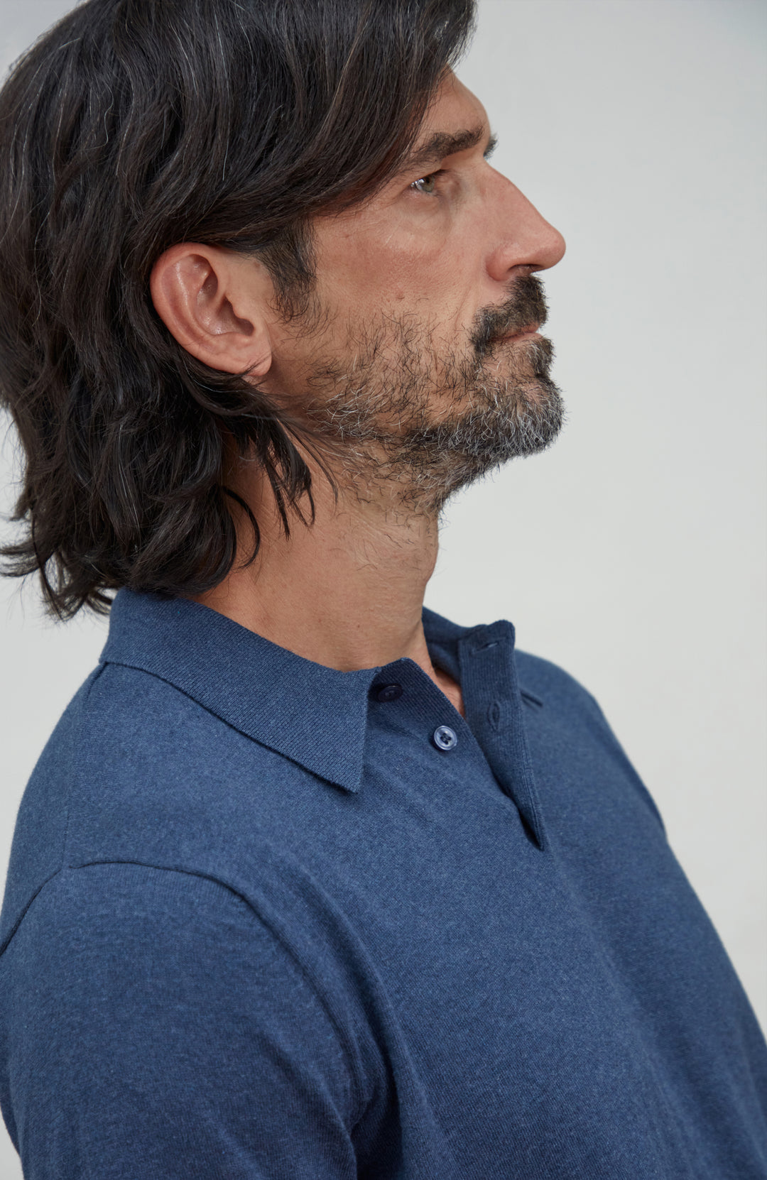A man in profile wearing a dark cotton blue heathered polo shirt against a plain light gray background. Made by Paul James Knitwear.