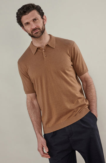 A man wearing a cottton short-sleeve knit polo shirt in camel brown by Paul James Knitwear, paired with dark navy trousers, standing against a plain light beige studio background.