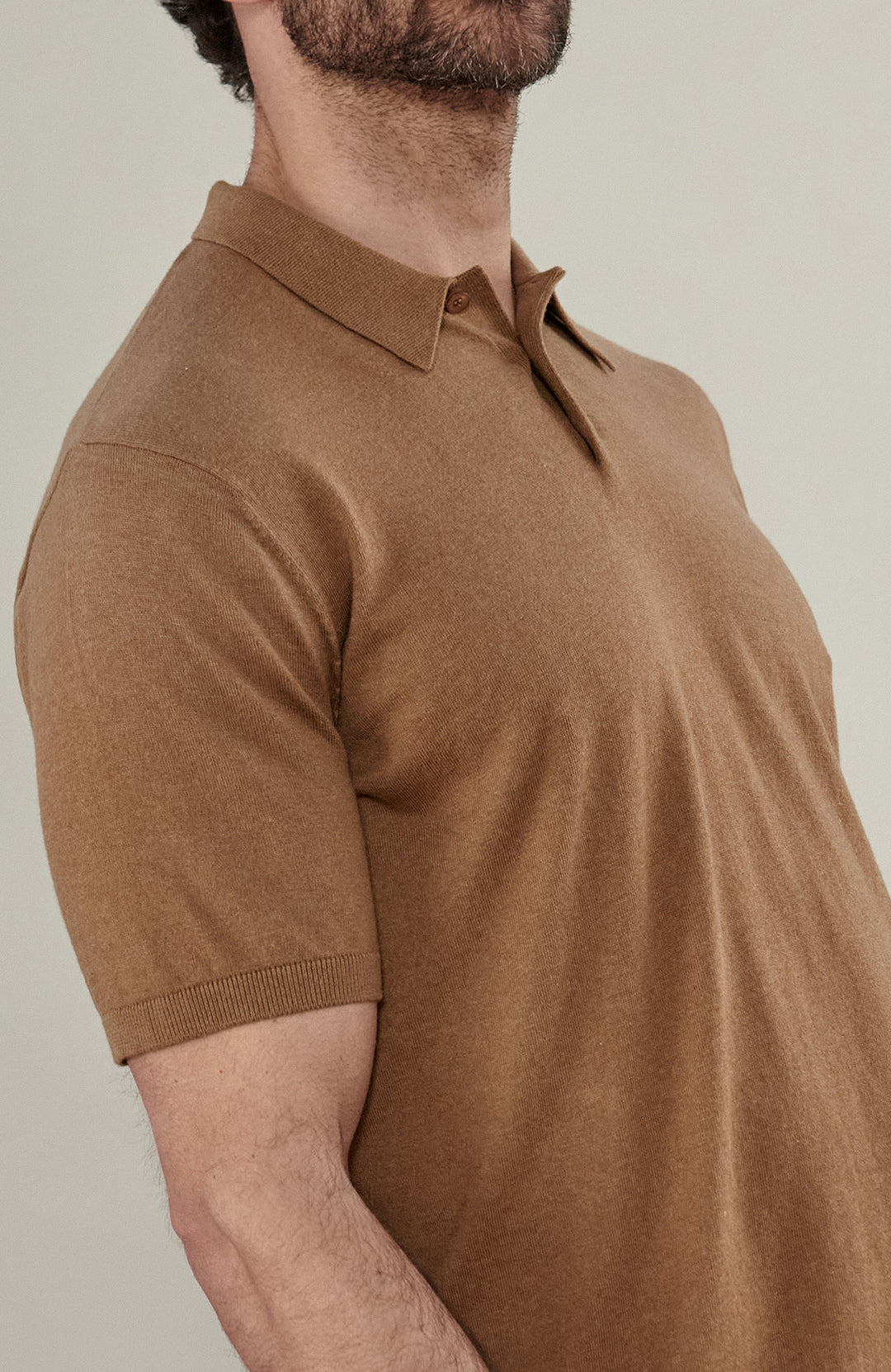 A man wearing a cottton short-sleeve knit polo shirt in camel brown by Paul James Knitwear, paired with dark navy trousers, standing against a plain light beige studio background.