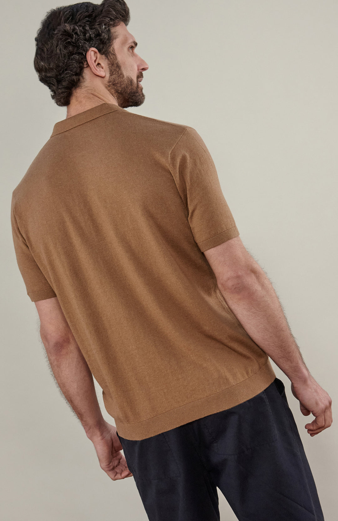 A man wearing a cottton short-sleeve knit polo shirt in camel brown by Paul James Knitwear, paired with dark navy trousers, standing against a plain light beige studio background.