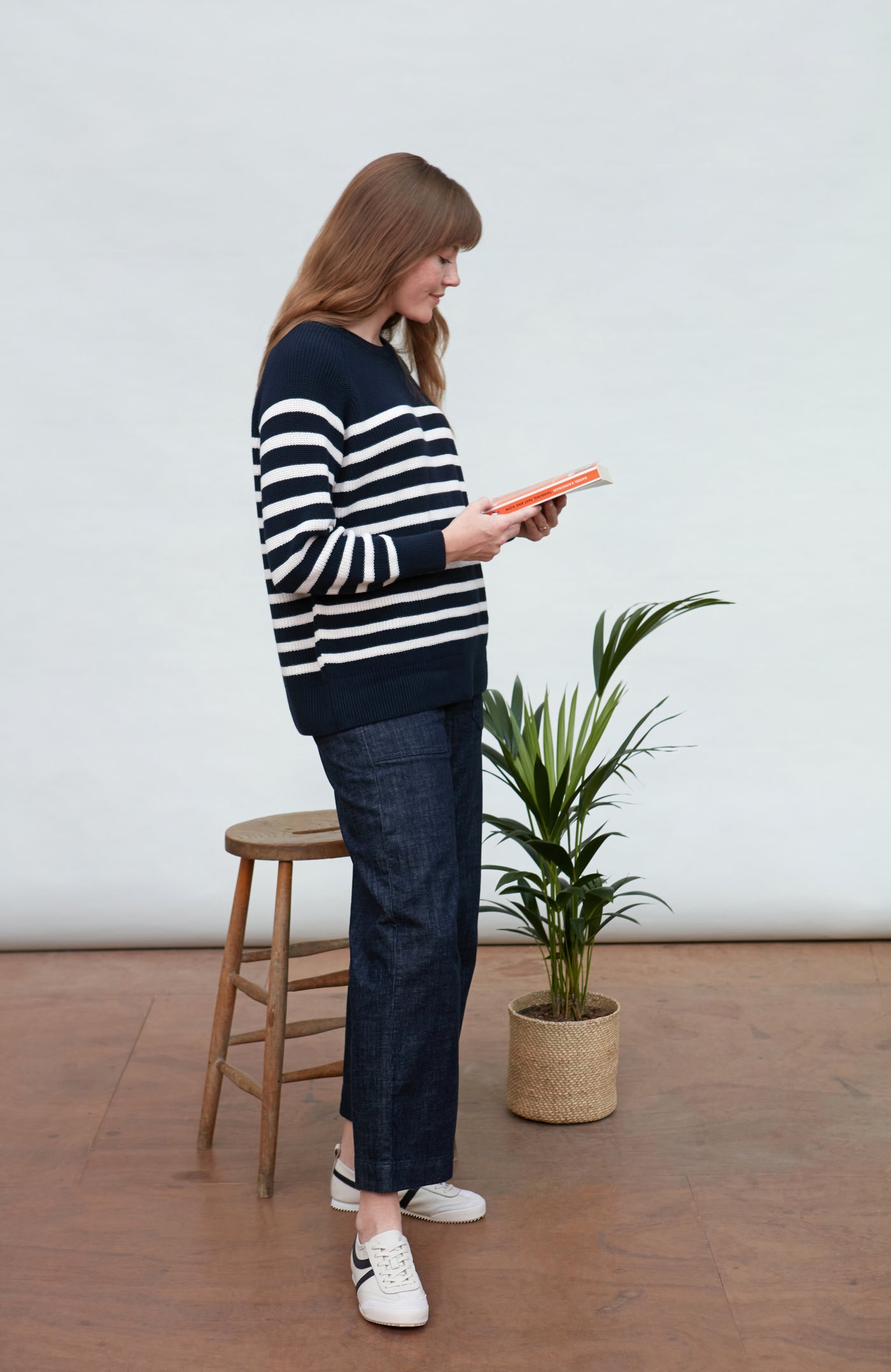 Woman in a striped breton sweater and jeans standing next to a plant in a minimalistic room.