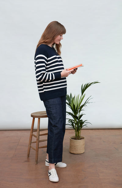 Woman in a striped breton sweater and jeans standing next to a plant in a minimalistic room.