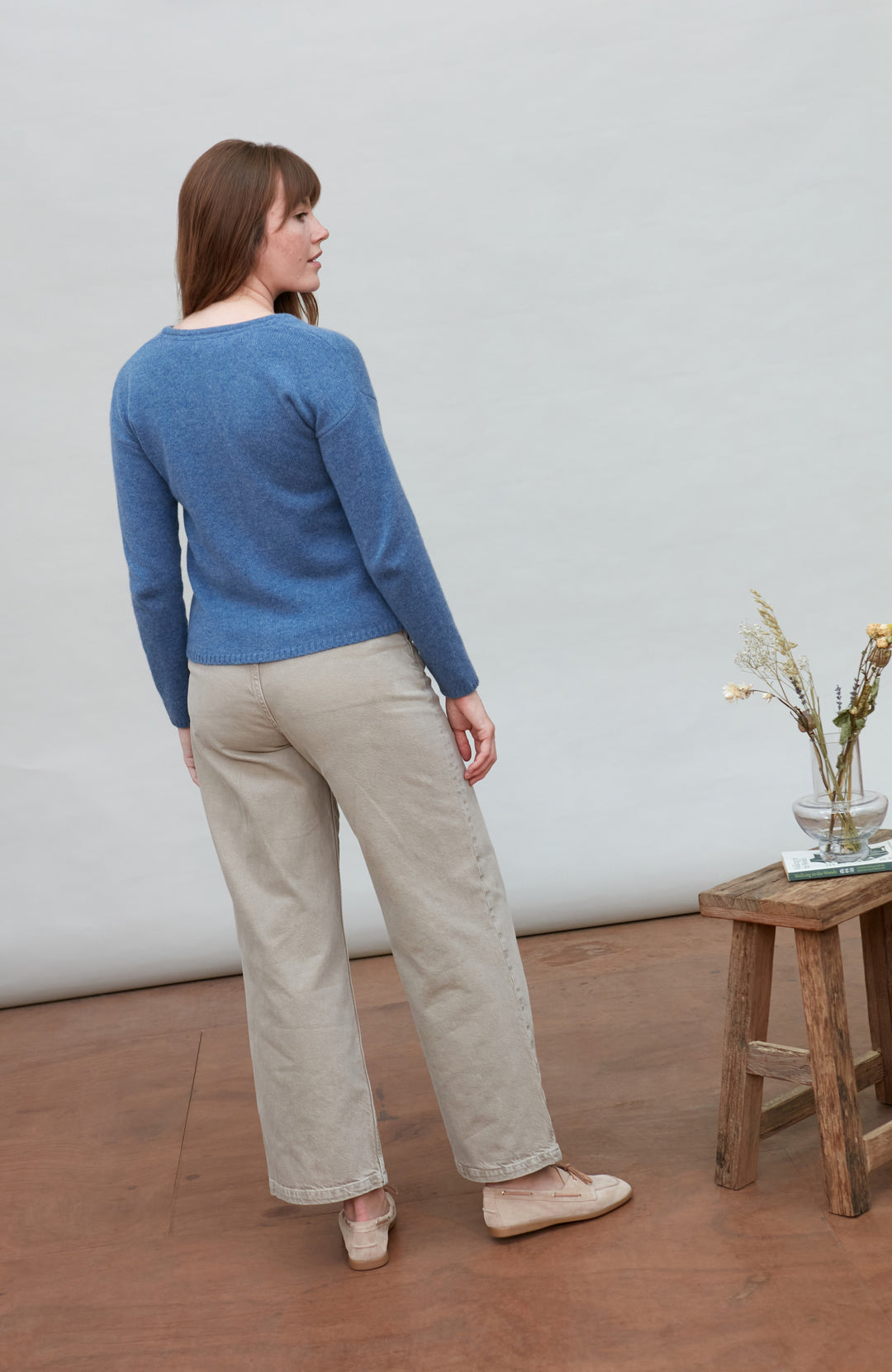 Woman wearing a blue wool sweater and beige pants standing in a room with a wooden floor and white wall.