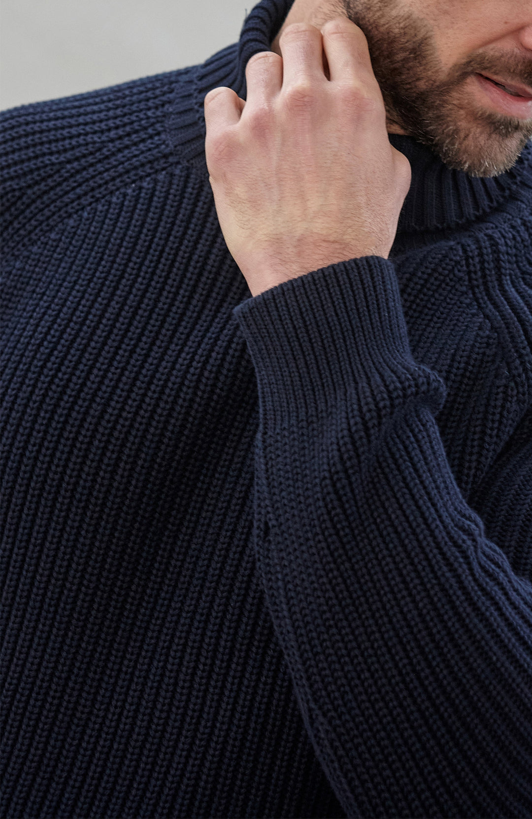 Man wearing a dark blue ribbed cotton roll neck sweater by Paul James Knitwear and brown pants holding an umbrella against a plain background