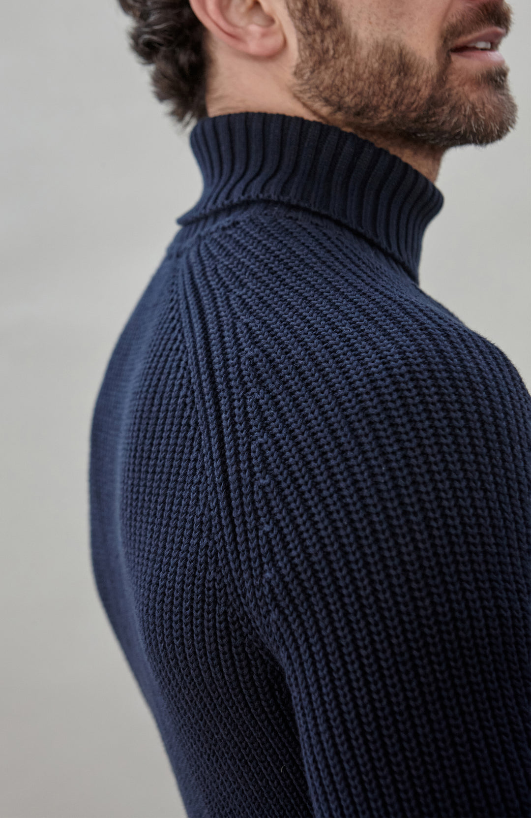 Man wearing a dark blue ribbed cotton roll neck sweater by Paul James Knitwear and brown pants holding an umbrella against a plain background